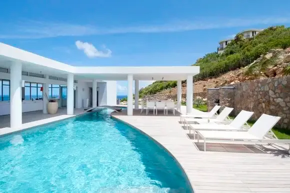 The modern outdoor living space centers on a curved pool with white lounge chairs on warm wood decking, a covered dining pavilion, and natural stone accents.                                                                                                                                                                                                                                                                                                                                                              +
 