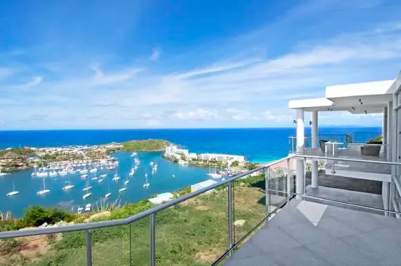 Glass railings frame an elevated terrace with sweeping views of the harbor, where dozens of sailboats anchor against a backdrop of tropical coastline and endless blue sky.                                                                                                                                                                                                                                                                                                                                                +
 