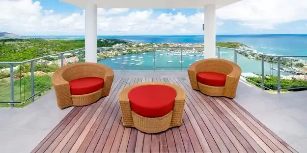 Elegant wicker seating with vibrant red cushions invites relaxation on the wooden deck, while sailboats anchor in the marina and the ocean stretches to the horizon.                                                                                                                                                                                                                                                                                                                                                     +
 