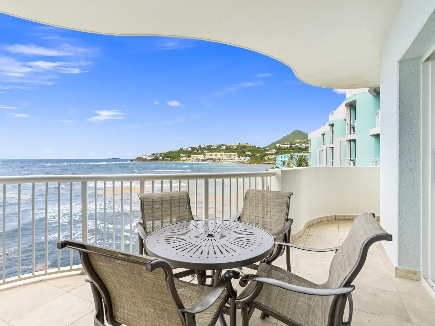Balcony with round metal table and four chairs overlooking ocean, coastal town, green hills, and blue sky