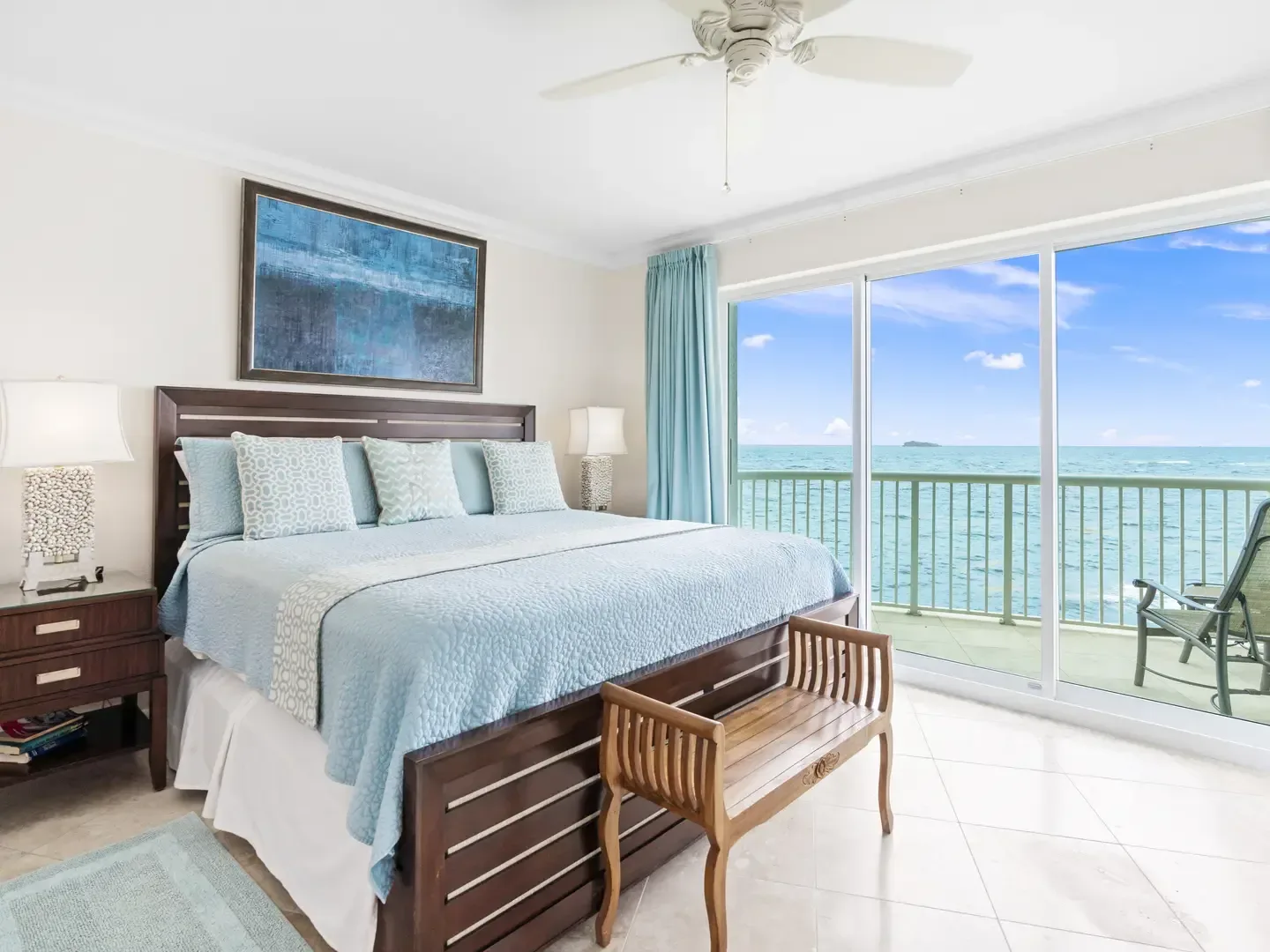 Master bedroom with ocean view, large bed, blue bedding, wooden furniture, and sliding glass doors leading to a balcony overlooking the sea