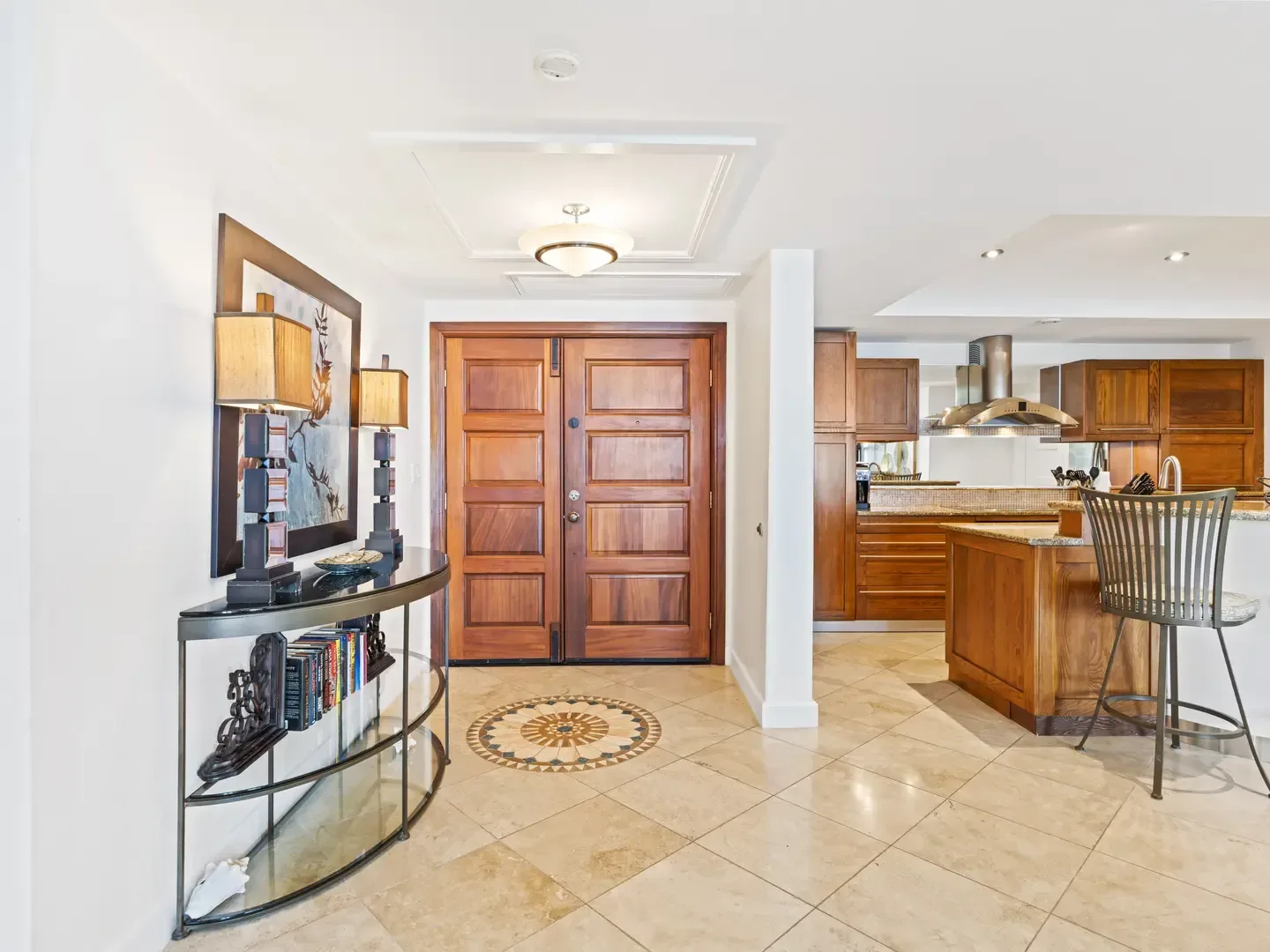 Luxury villa entrance with wooden double doors, glass console table, and open kitchen with dark wood cabinets and granite island