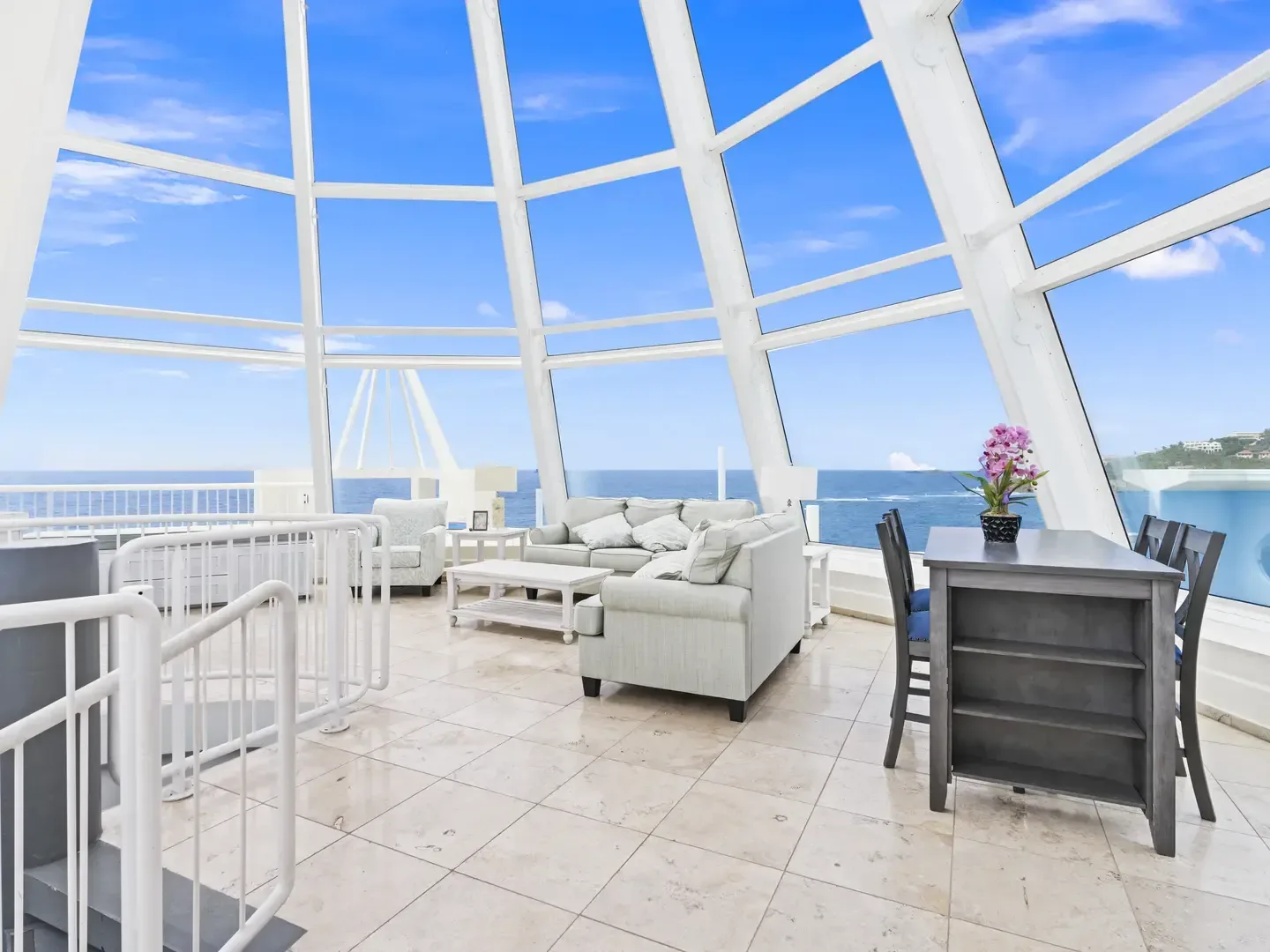 Bright open-concept living and dining space with panoramic ocean views through glass dome windows, light gray sofa, dining table with orchids