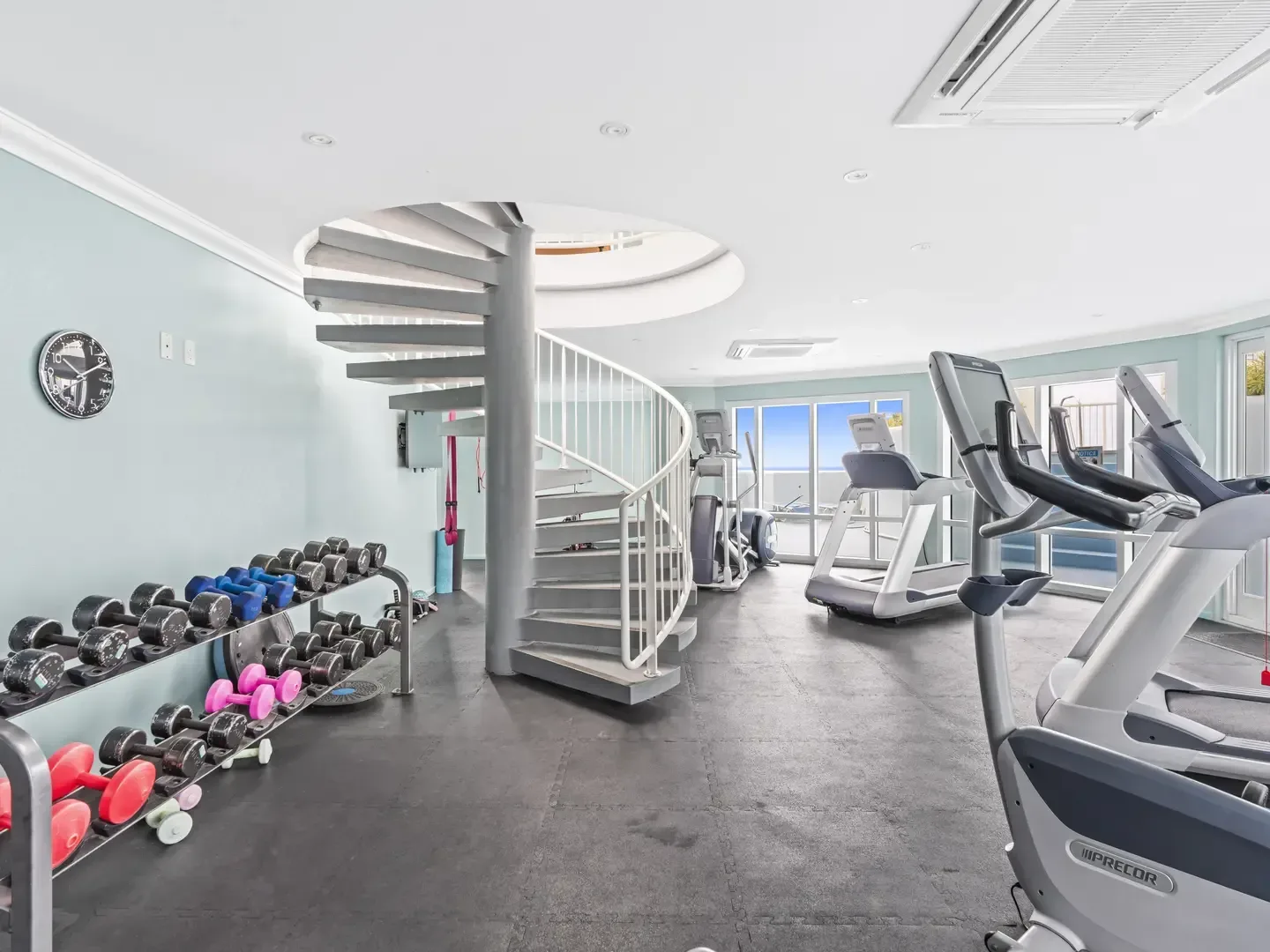 Modern villa gym with dumbbells, Precor exercise machines, spiral staircase, and ocean view through large windows