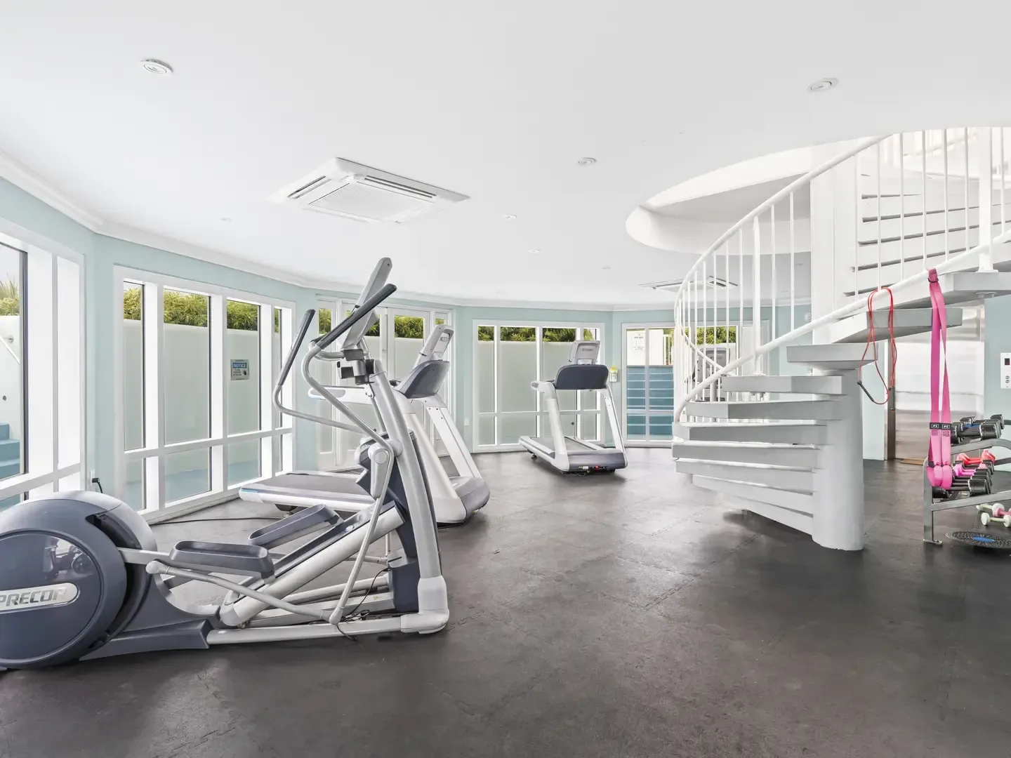Modern gym with Precor ellipticals, treadmill, weights, white spiral staircase, and large windows overlooking greenery