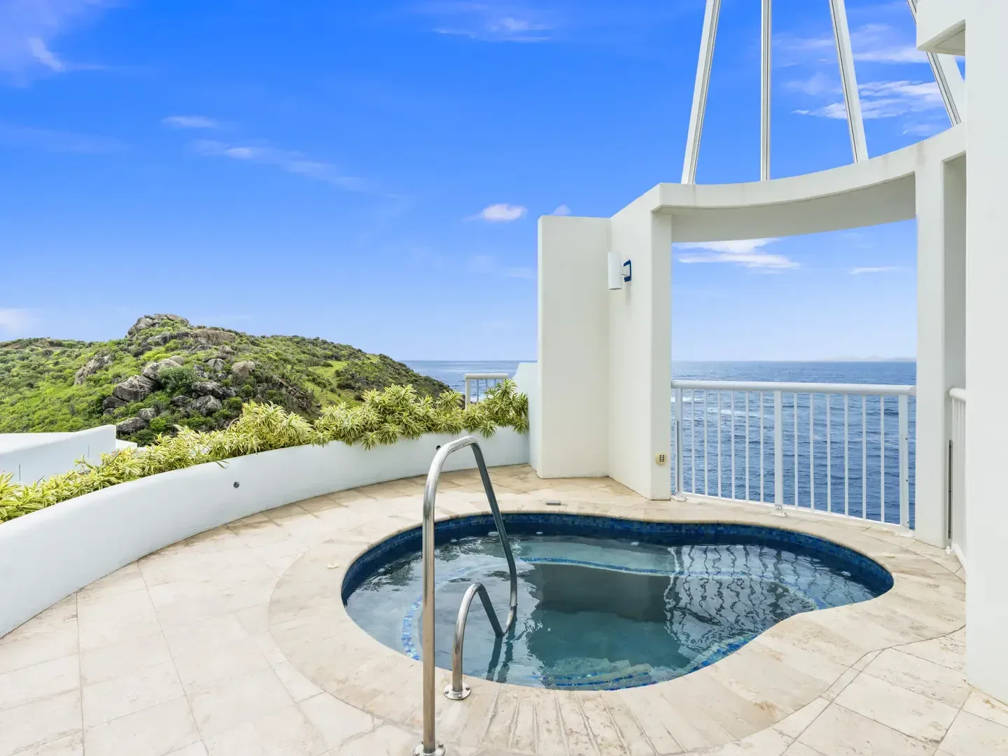 Circular outdoor jacuzzi with stainless steel ladder, ocean and green rocky hill view, white villa architecture, bright blue sky