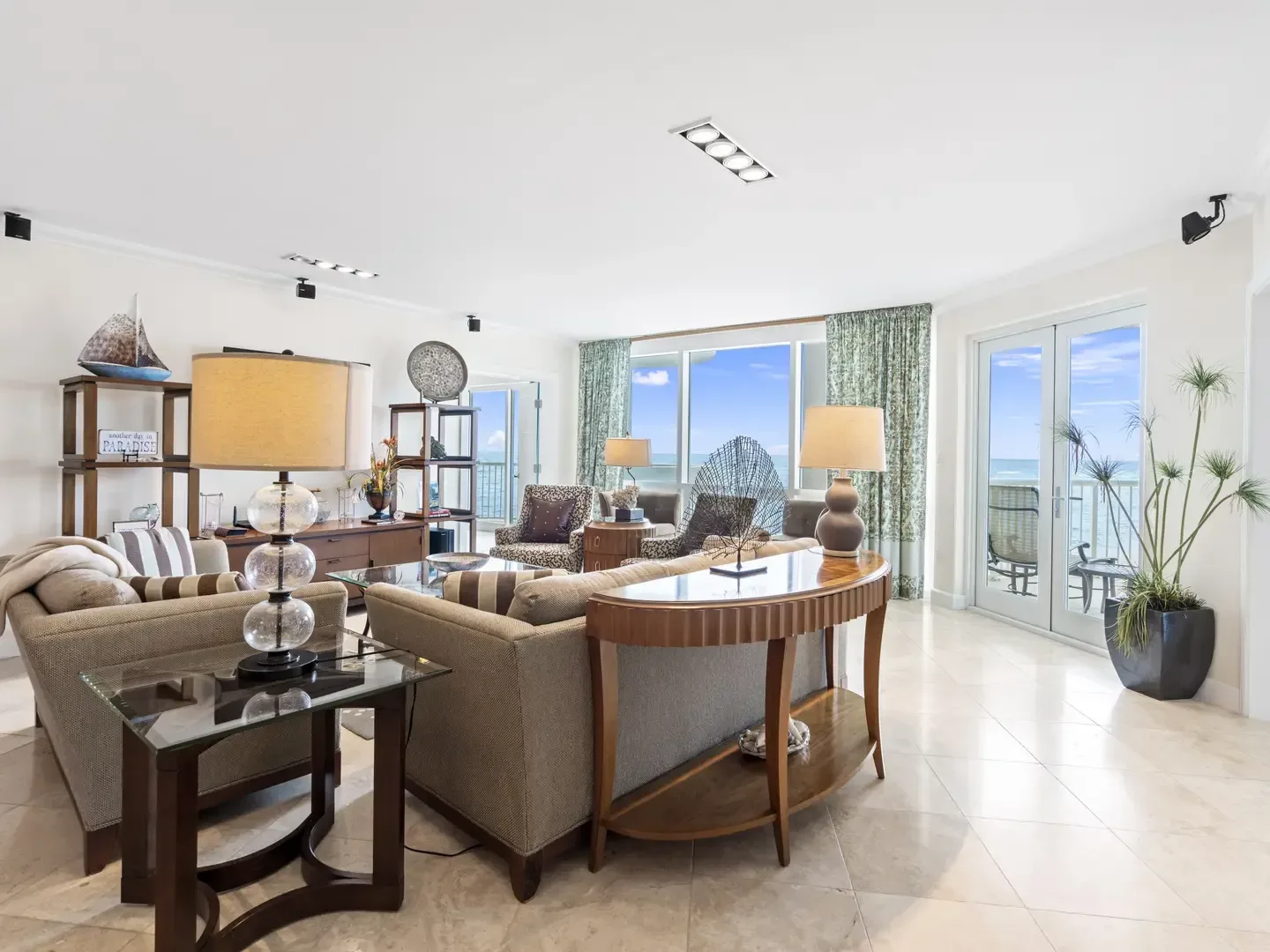 Spacious villa living room with beige sofas, glass tables, ocean views through large windows, and coastal decor
