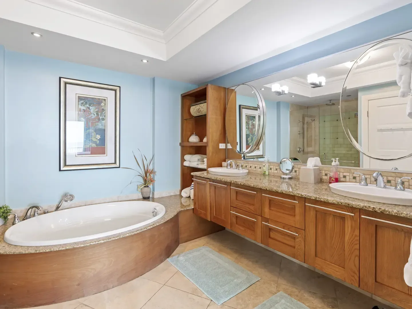 Spacious luxury bathroom with light blue walls, wooden cabinets, double sinks, oval bathtub, large mirrors, and granite countertops
