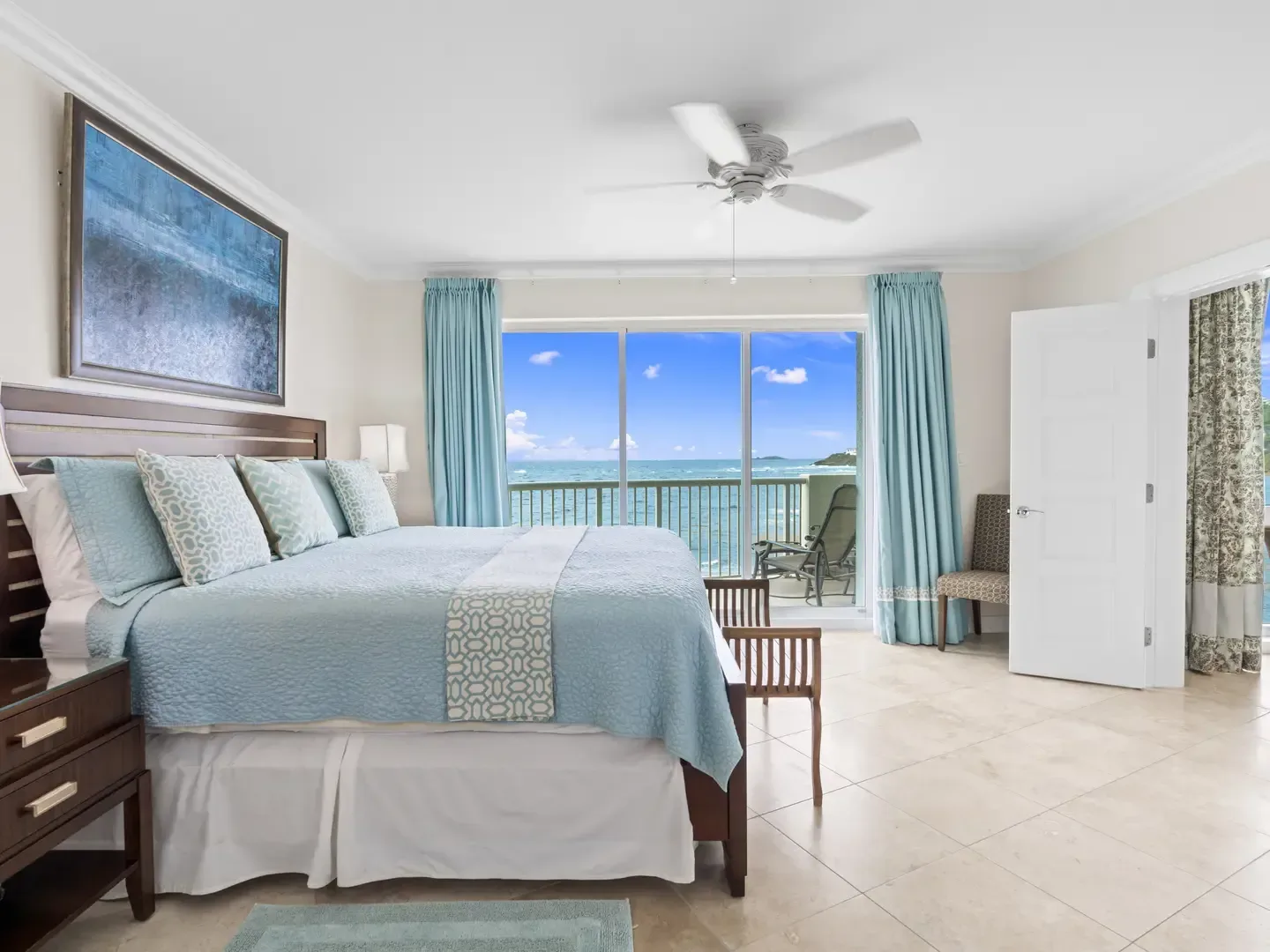 Spacious master bedroom with king bed, light blue bedding, ocean view through sliding glass doors, ceiling fan, and balcony with lounge chairs