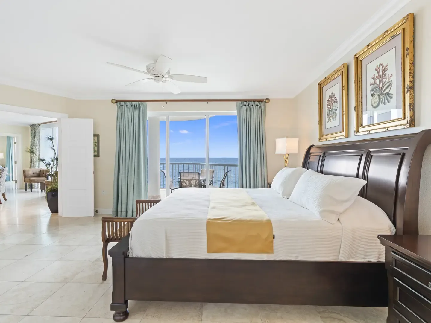 Spacious luxury bedroom with dark wood bed, white linens, gold throw, ocean view, and coral-themed artwork