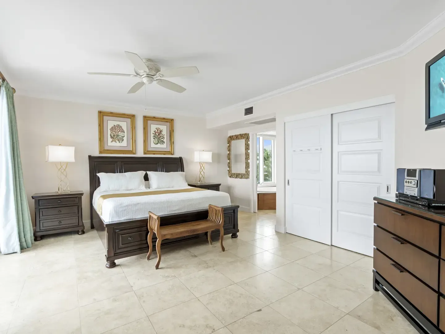 Spacious master bedroom with dark wood king-size bed, white linens, gold accents, wooden bench, and view of tropical greenery