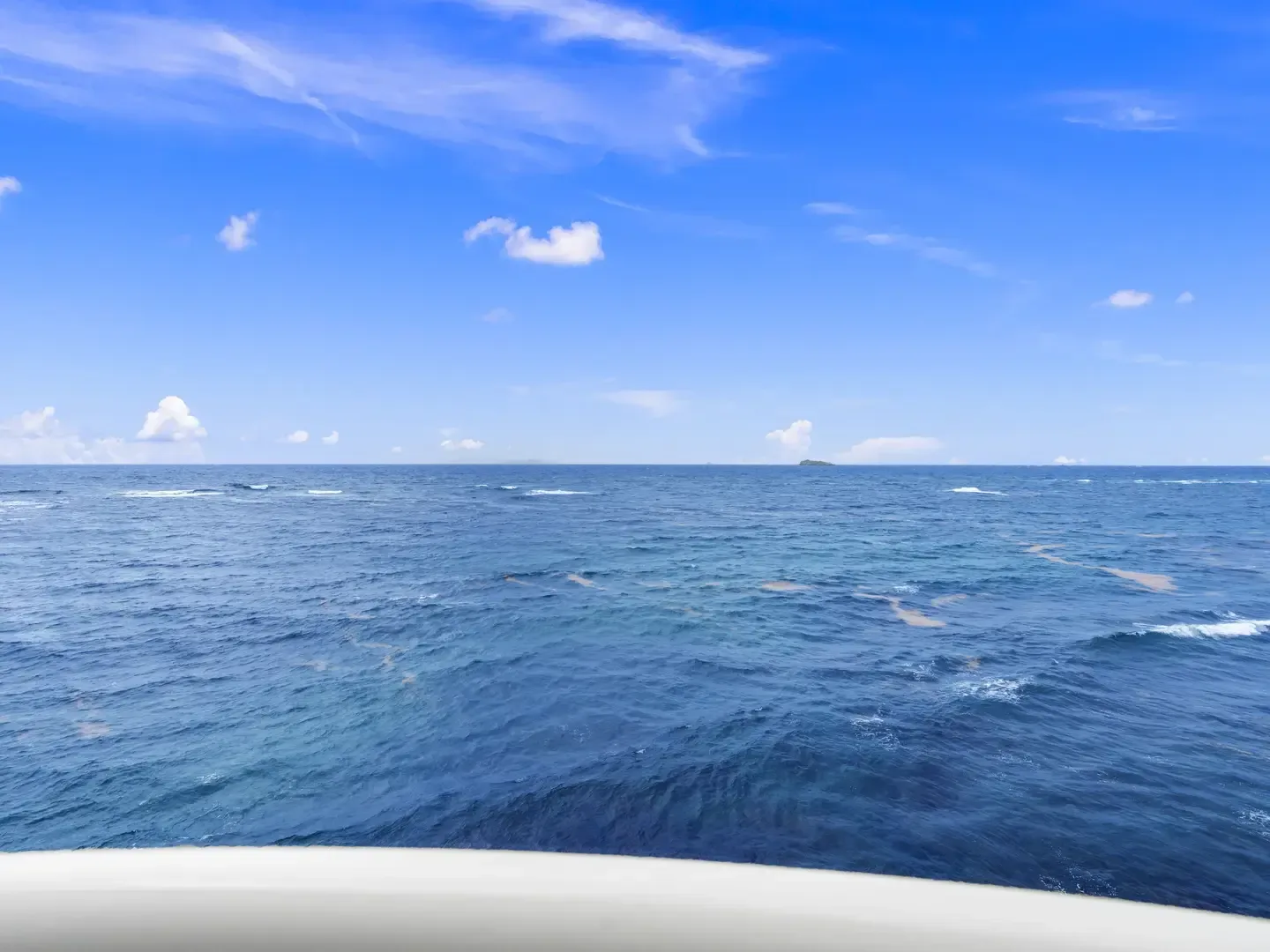 Panoramic ocean view with deep blue waters, scattered white clouds, and a distant island, viewed from a white railing