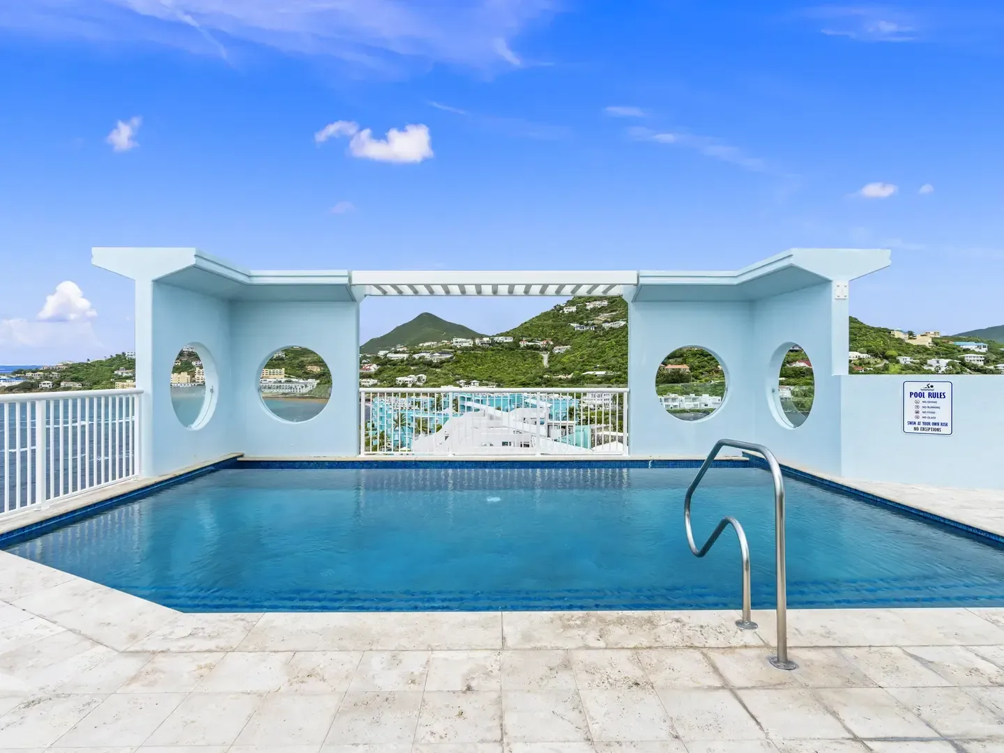 Luxury outdoor pool with blue water, stone tiles, scenic ocean and hill views, light blue structure with circular cutouts
