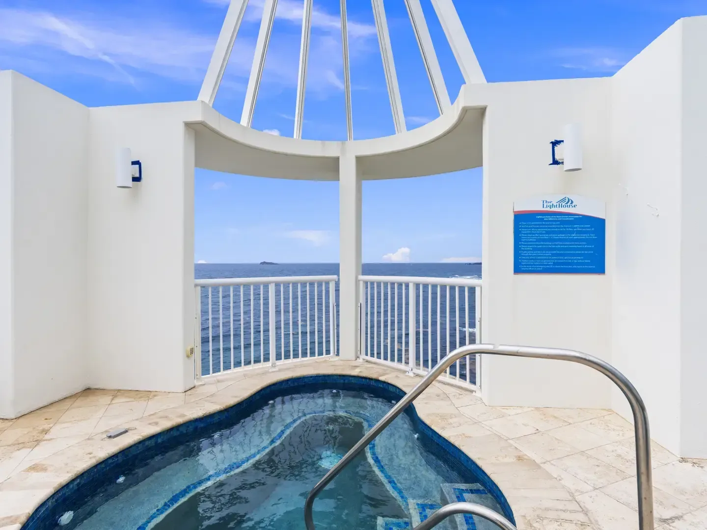 Small pool with blue tiles and metal handrail on stone deck, white arched walls, ocean view with distant island, and The LightHouse sign