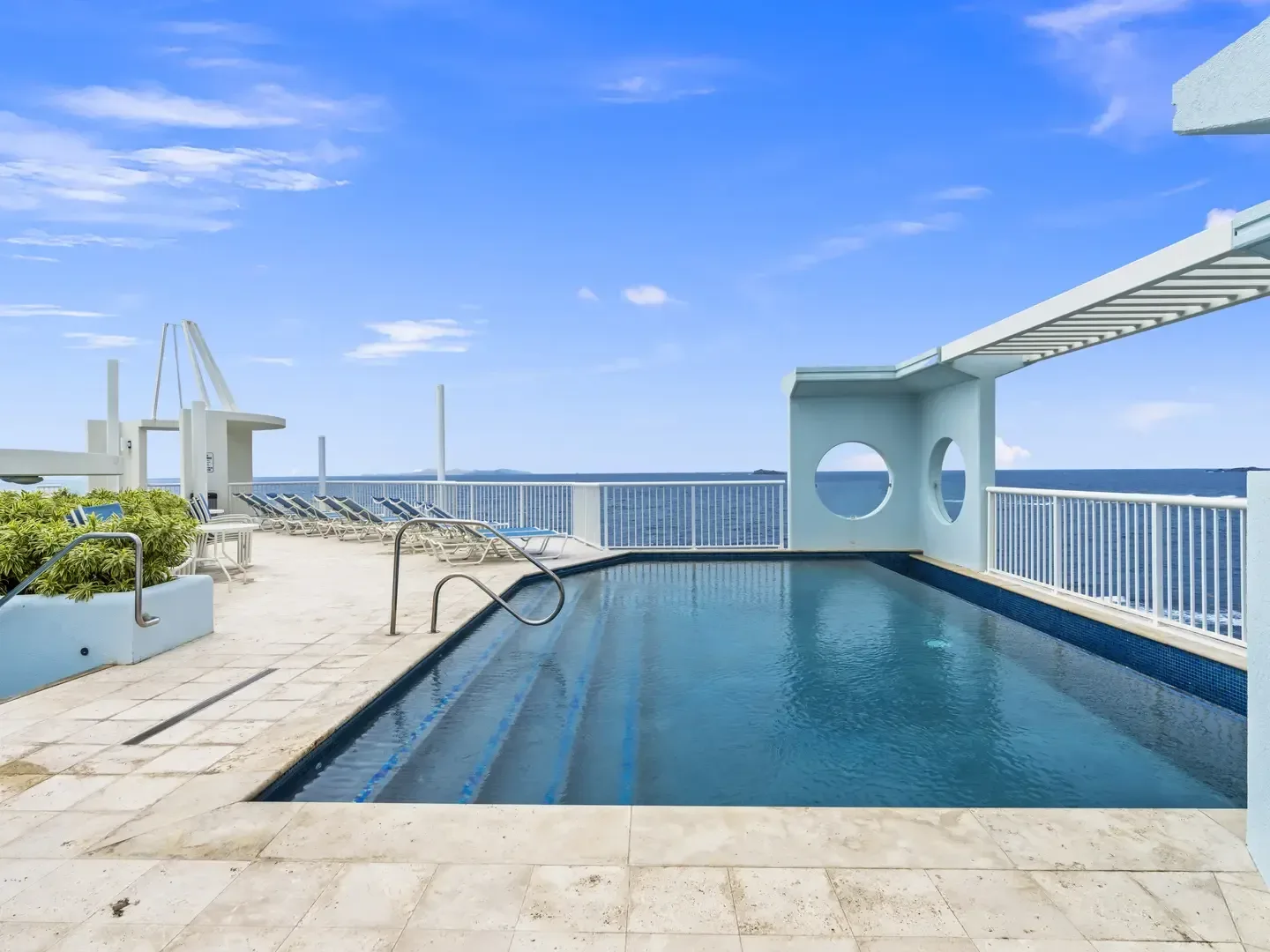 Luxury outdoor pool with ocean view, white lounge chairs, circular windows, and blue sky