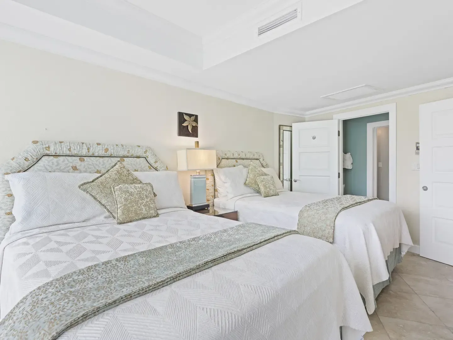 Spacious bedroom with two neatly made beds, white geometric-patterned linens, decorative pillows, and turquoise bathroom visible