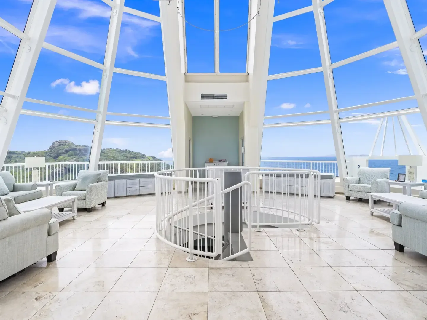 Interior of lighthouse villa with spiral staircase, large glass windows, light blue furniture, and ocean-mountain views