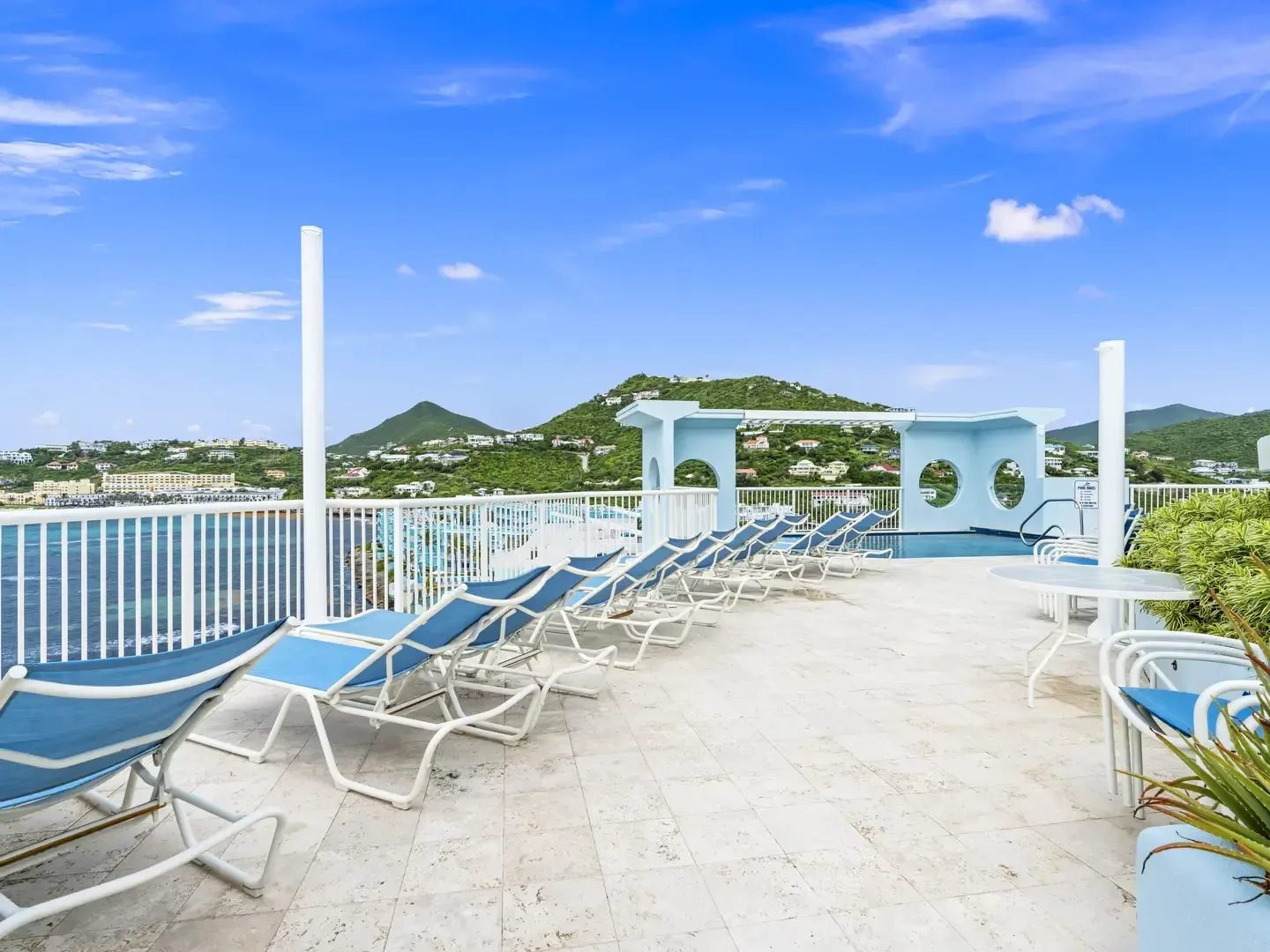 Outdoor terrace with blue lounge chairs, white railings, ocean and hill views, light blue architectural structure, and clear blue sky