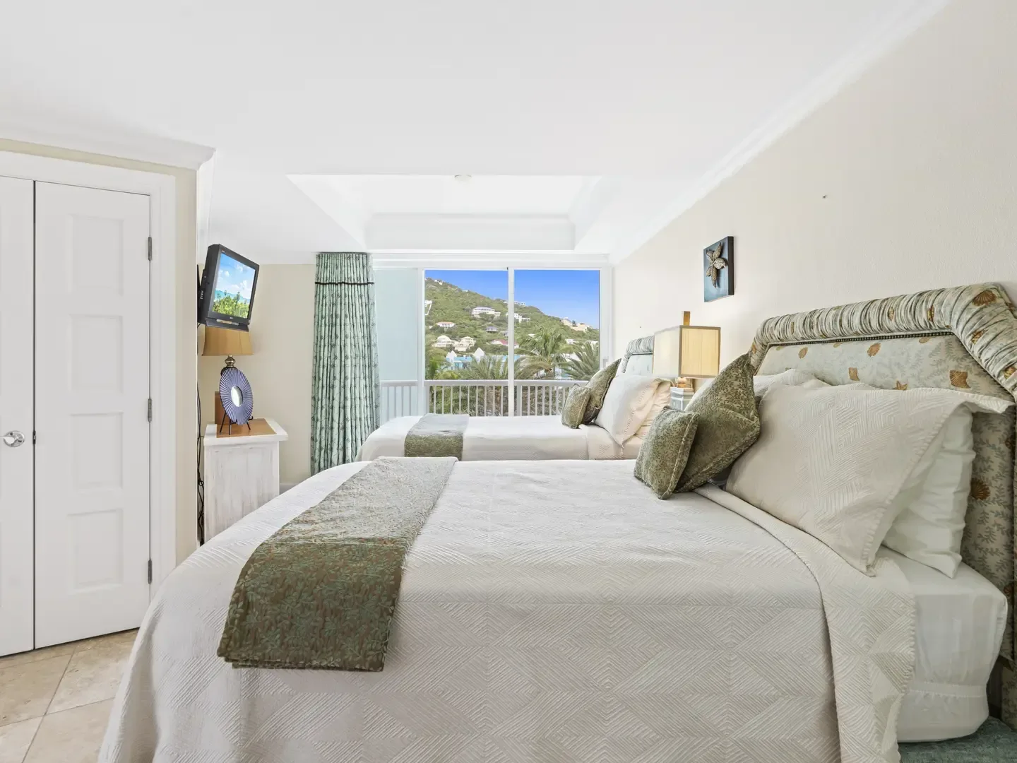 Bright bedroom with two beds, white and green bedding, sliding glass doors to a balcony with mountain and house views