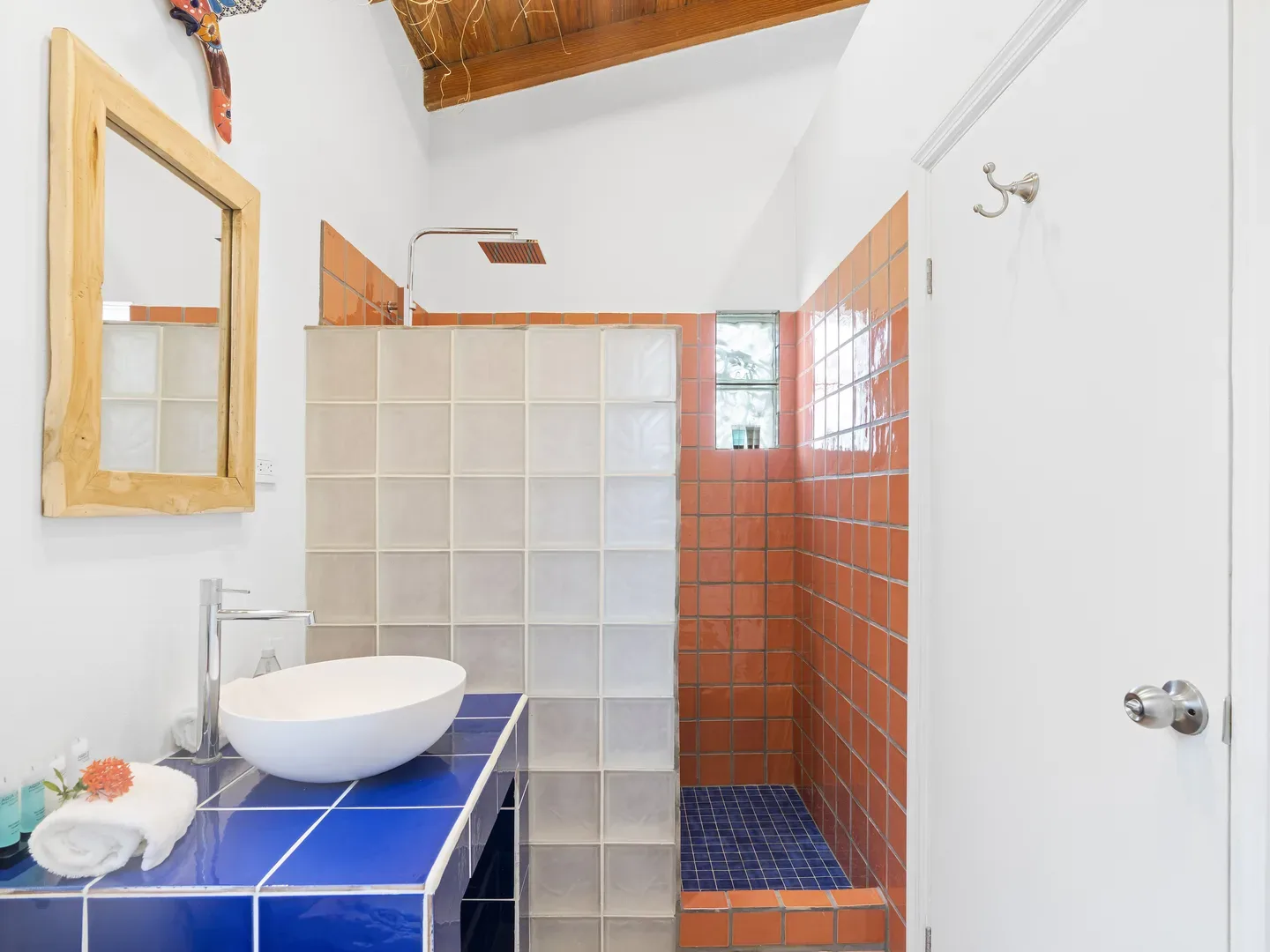 Modern bathroom with blue and orange tile accents, white vessel sink, wooden mirror frame, and glass block wall separating shower area