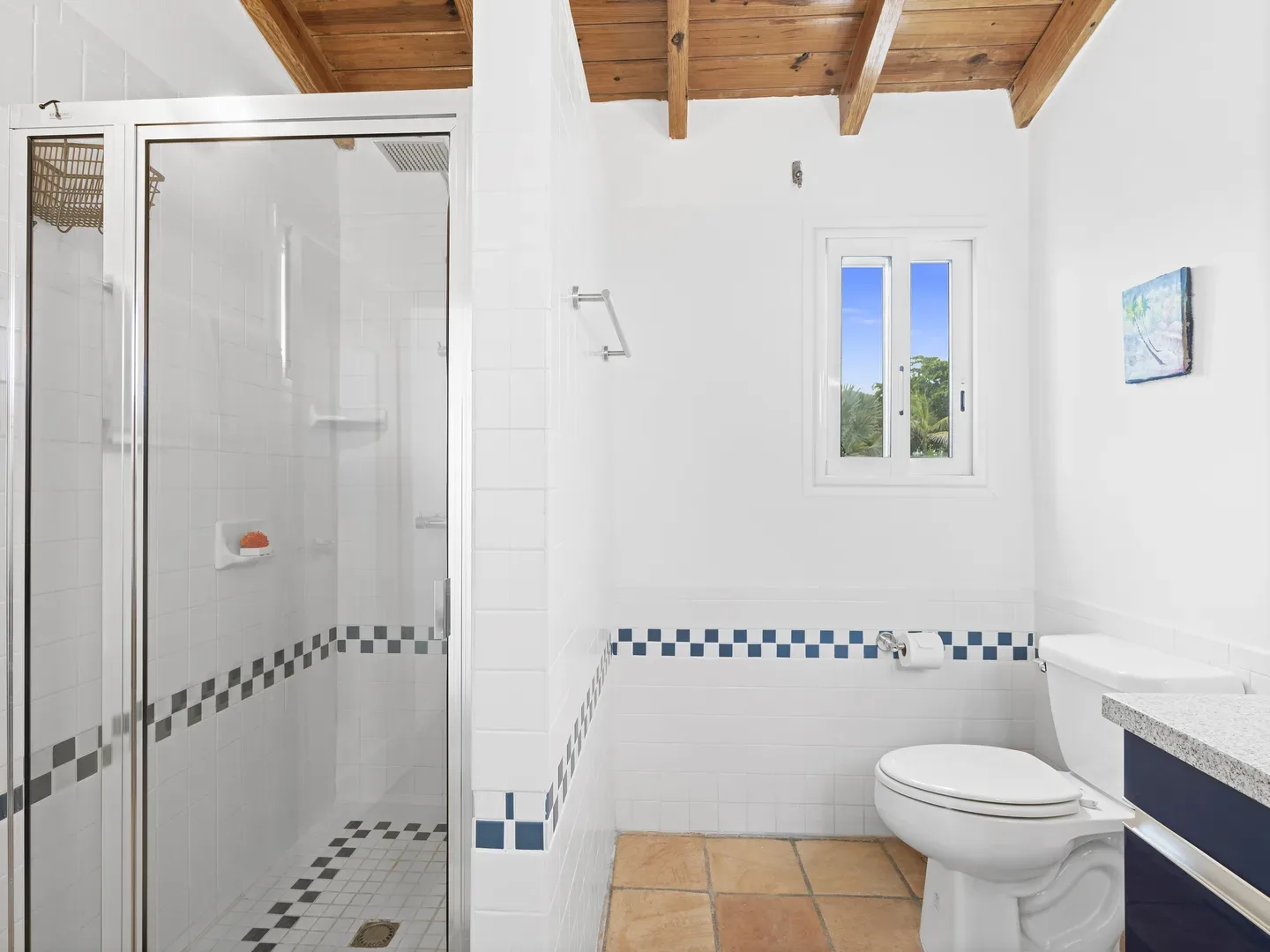 Bright modern bathroom with glass-enclosed shower, white toilet, granite countertop sink, terracotta floor tiles, blue-accented white tile walls, wooden beam ceiling, and window showing tropical greenery and blue sky