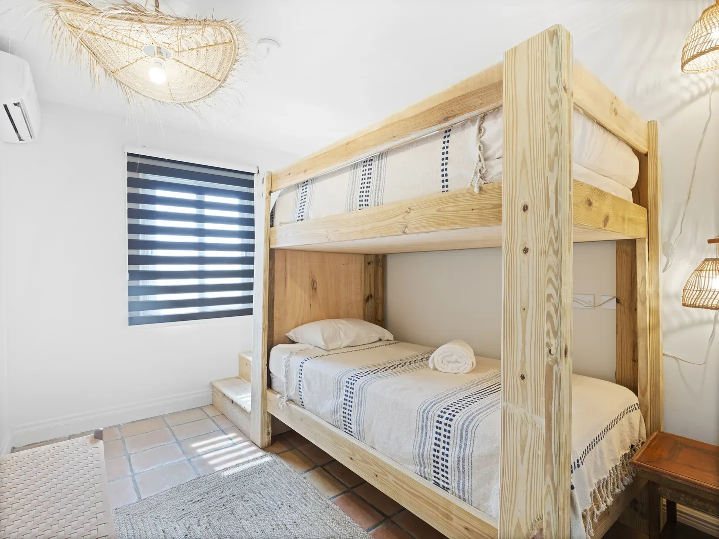 Bright bedroom with wooden bunk bed, white and blue striped bedding, terracotta tile floor, wicker lighting, and window with dark blue blinds
