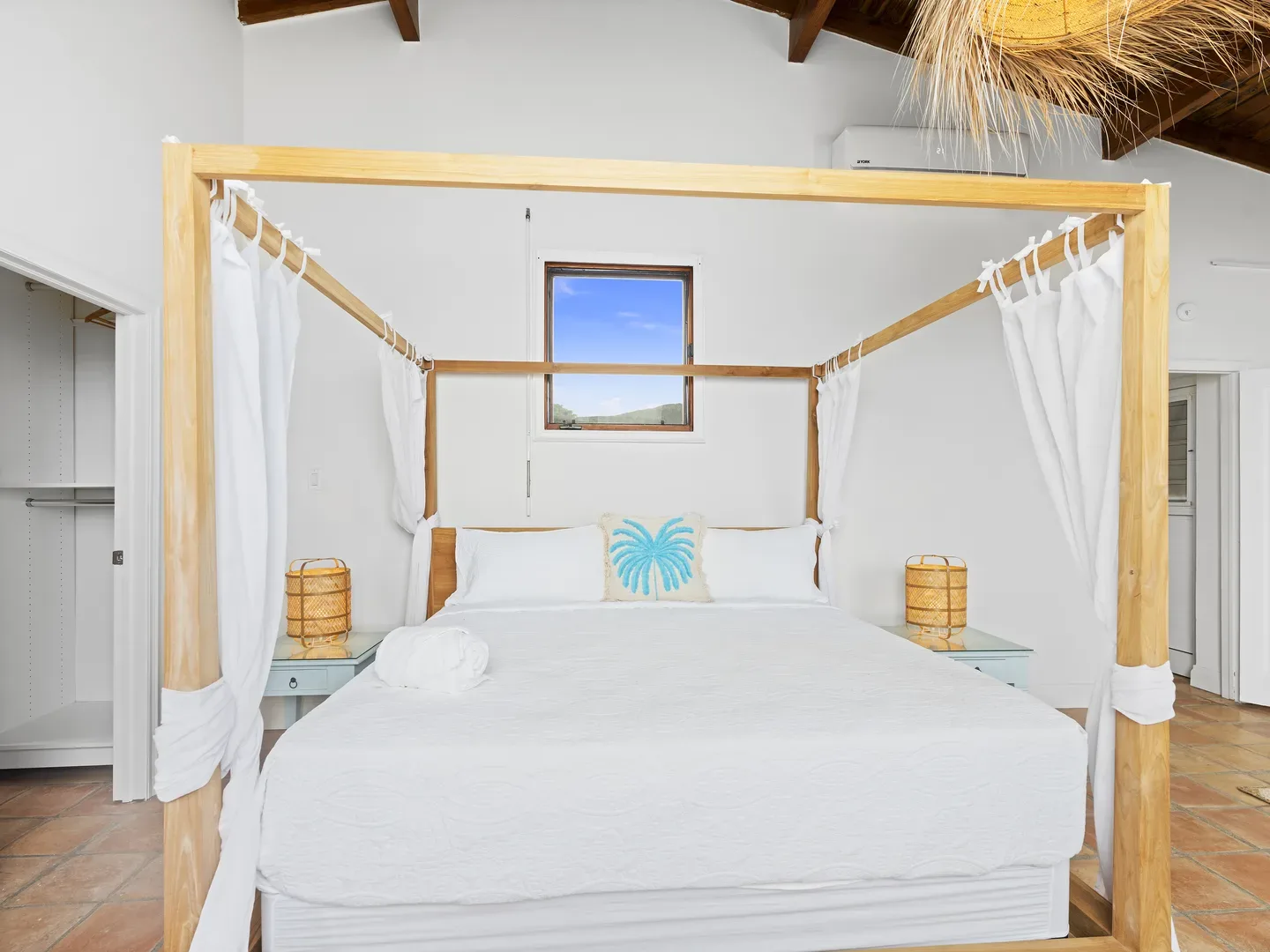 Bright bedroom with a wooden four-poster bed, white linens, turquoise palm pillow, wicker bedside lamps, and a window view of blue sky and green hills