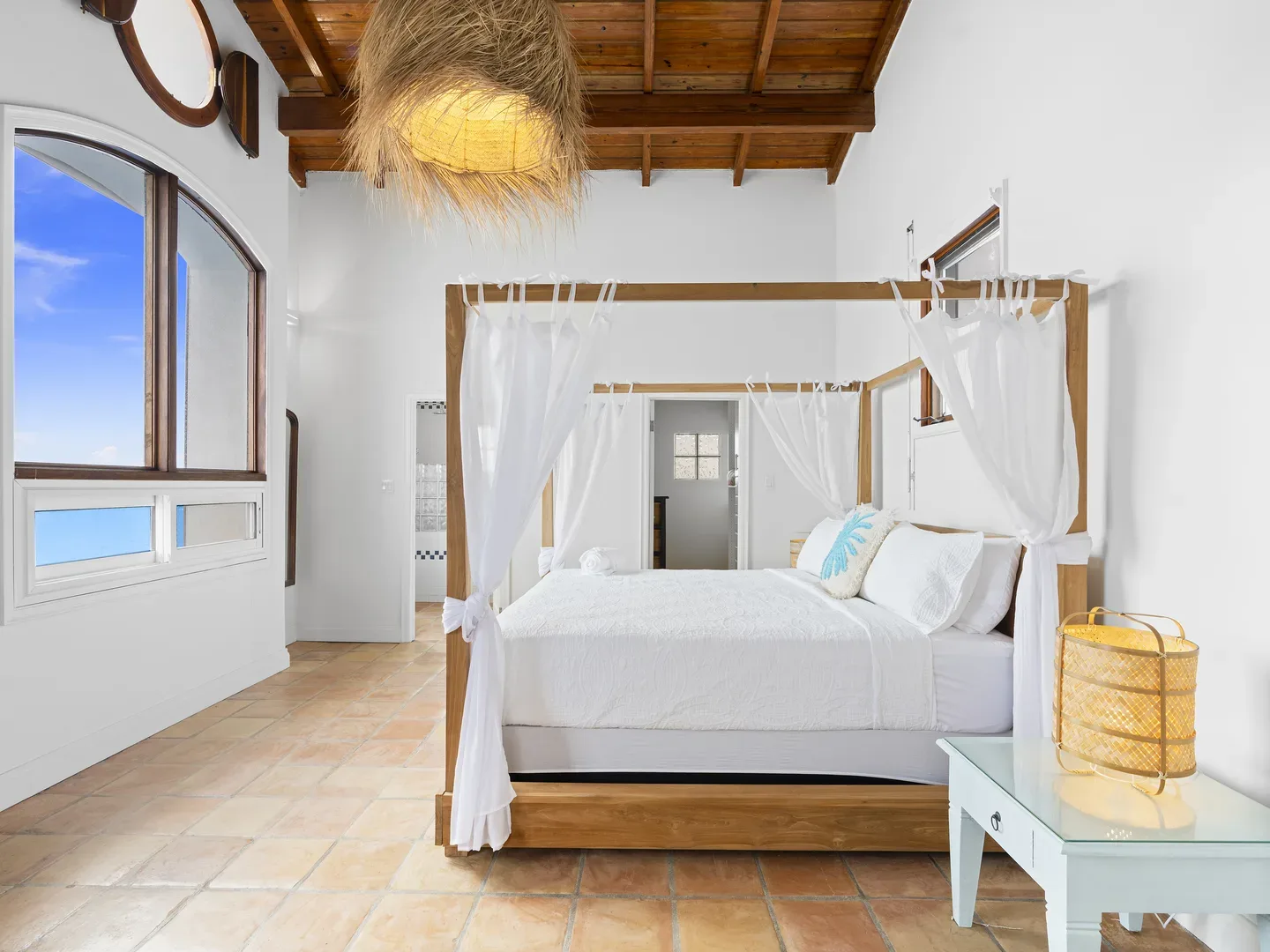 Luxury bedroom with four-poster bed, white linens, arched window showing blue sky, wooden ceiling beams, and woven lighting fixtures