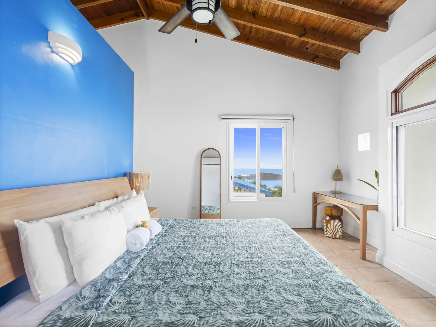 Spacious bedroom with king bed, blue accent wall, wooden ceiling beams, and ocean view through sliding window