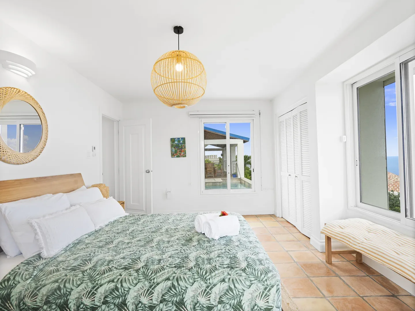 Bright tropical bedroom with large bed, green leaf-patterned duvet, wicker pendant light, and ocean view through windows