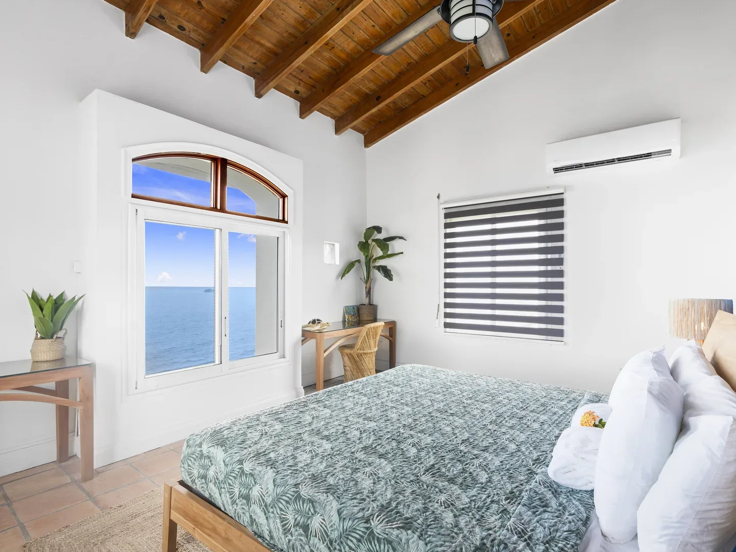 Bright tropical bedroom with wooden bed, green leaf-patterned bedspread, ocean view through arched window, exposed wood ceiling beams, and terracotta floor tiles
