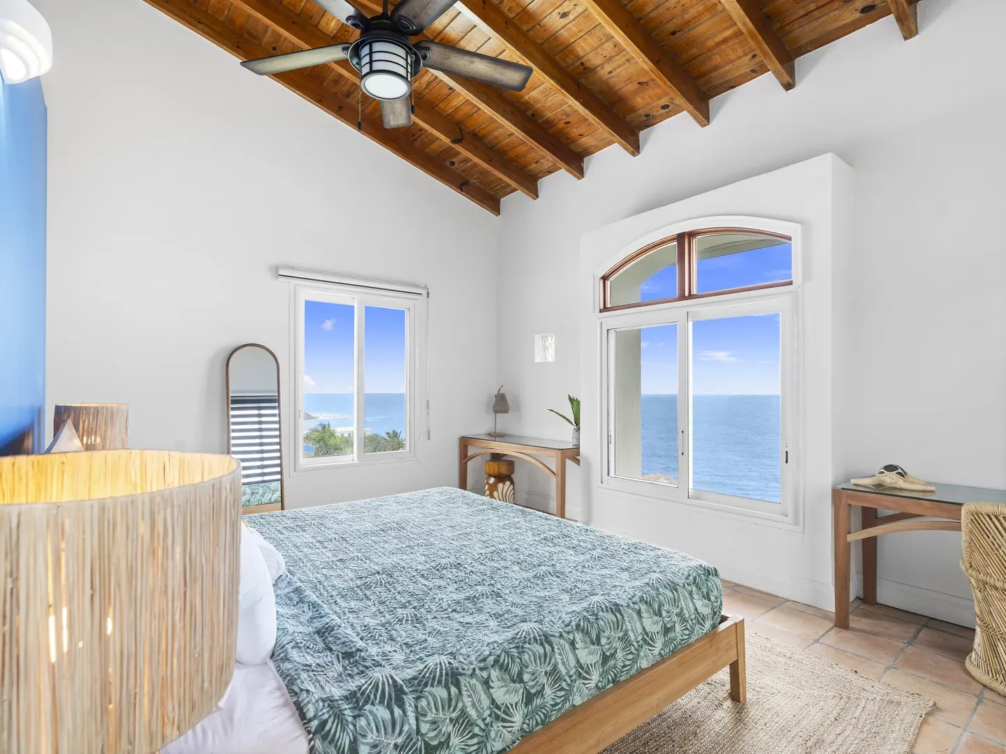 Bright bedroom with wooden ceiling beams, tropical-patterned bedspread, and panoramic ocean views through large arched window