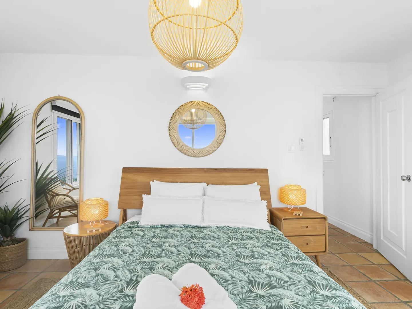 Tropical bedroom with ocean view through mirror, green leaf-patterned bedspread, wicker chandelier, and wooden furniture