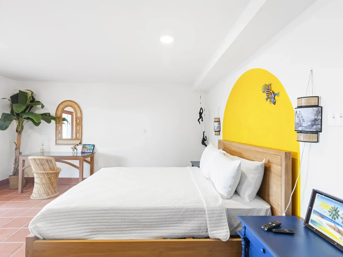 Spacious bedroom with white bedding, yellow arched accent wall, blue nightstand, wicker chair, and tropical decor