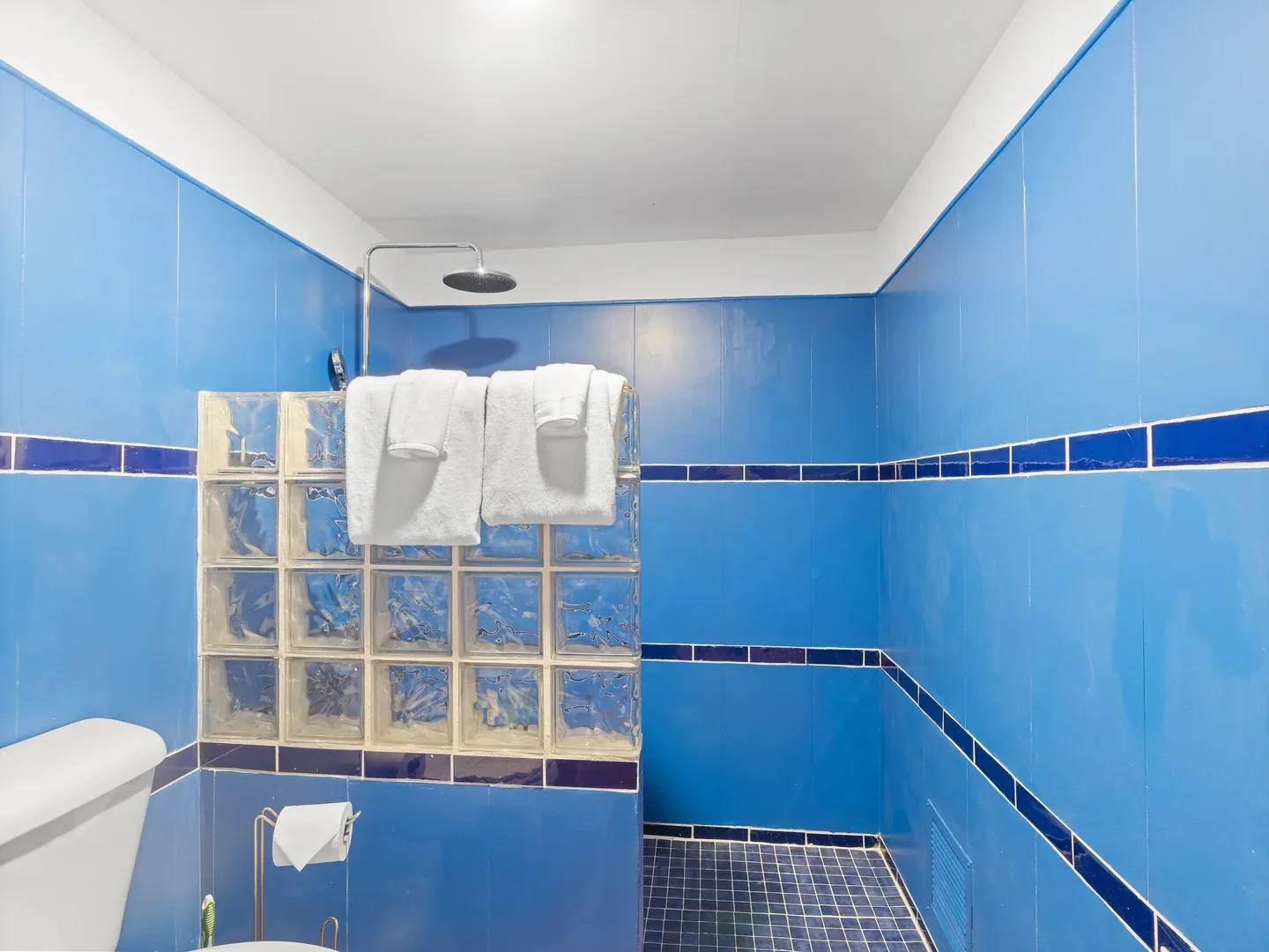 Bright blue-tiled bathroom interior with white ceiling, glass block partition, white towels, and rain showerhead