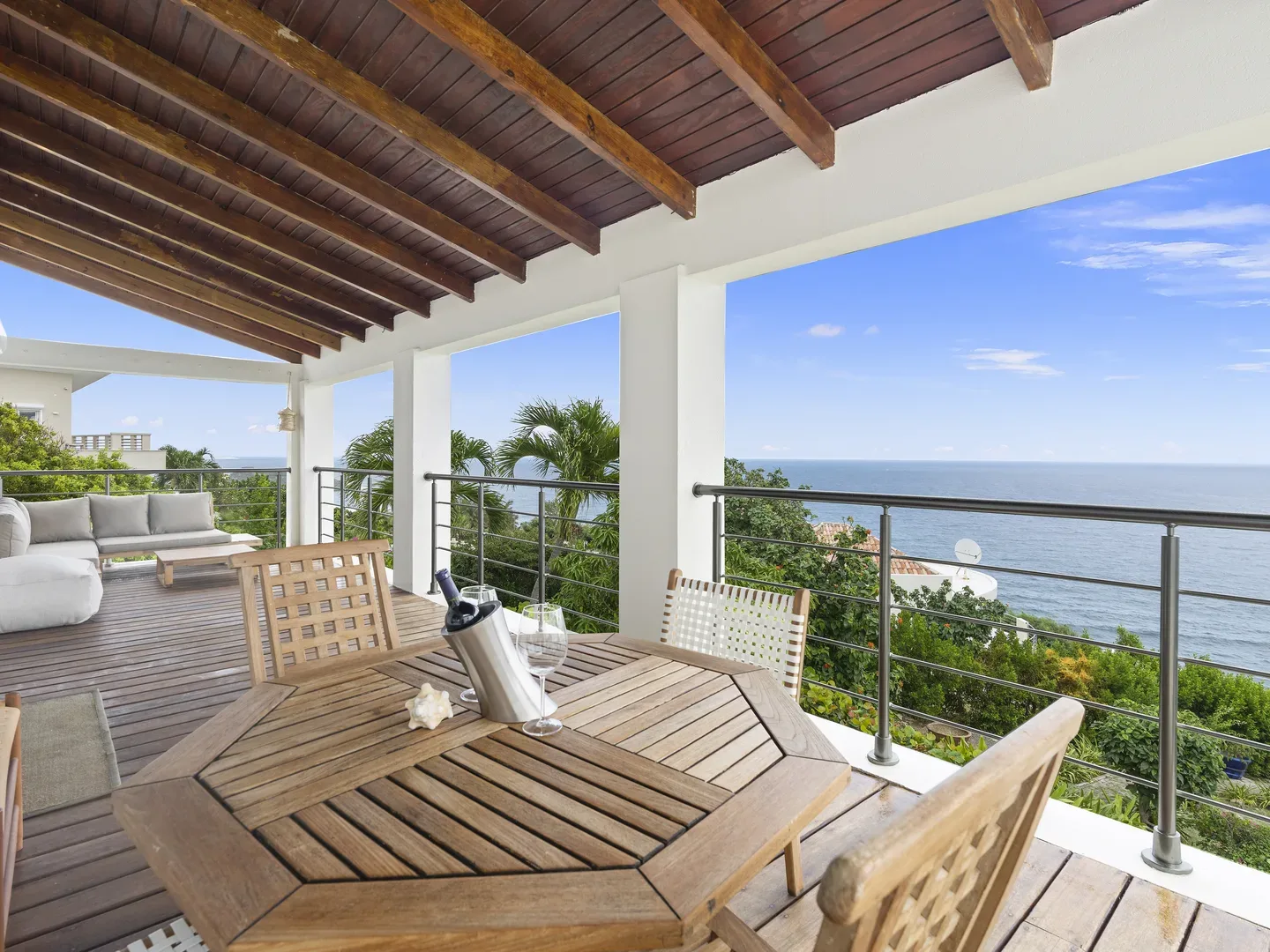 Covered wooden terrace with octagonal dining table, chairs, ocean view, gray sectional sofa, and metal railings
