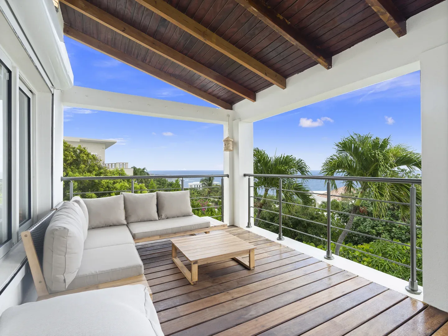 Covered terrace with beige sectional sofa, wooden coffee table, ocean view, and tropical greenery