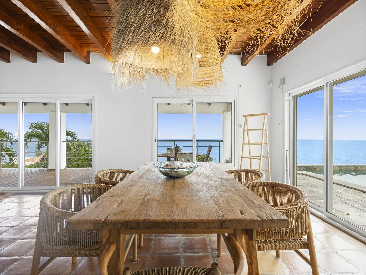 Rustic dining room with wooden table, wicker chairs, straw chandelier, and ocean view through glass doors