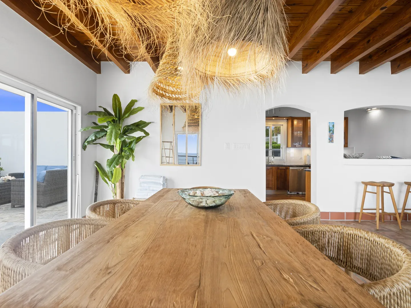 Spacious dining area with wooden table, wicker chairs, straw chandelier, sliding doors to patio with ocean views, and kitchen in background