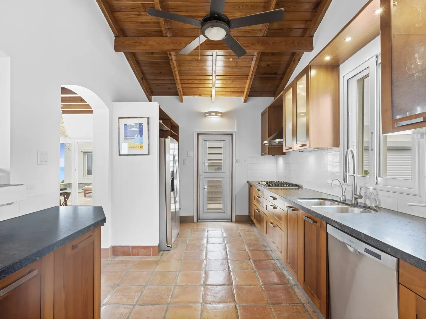 Modern kitchen with wooden cabinets, dark countertops, terracotta tile flooring, sea view through archway, exposed wooden ceiling beams, and ceiling fan
