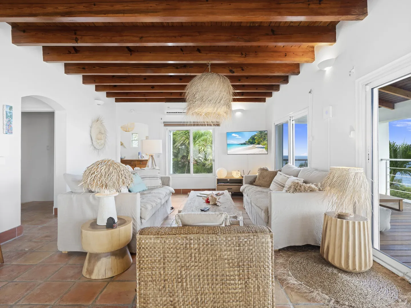 Bright living room with beige sofas, exposed wooden beams, straw pendant light, and ocean view through glass doors to a wooden terrace