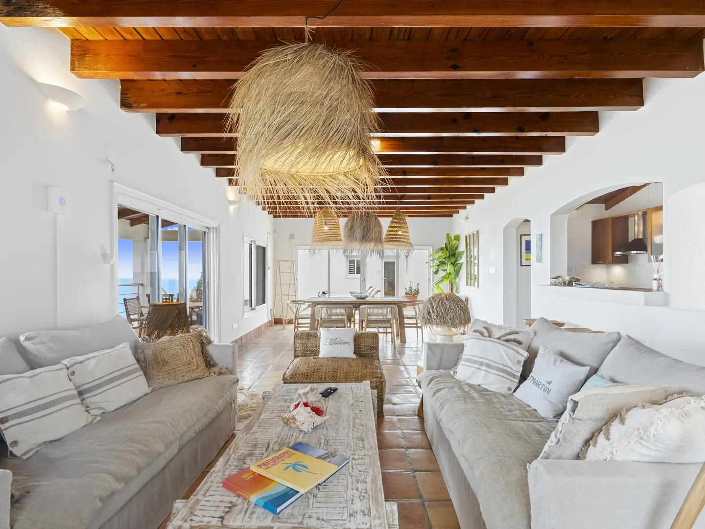 Spacious living room with gray sofas, wooden beams, straw pendant lights, and ocean view through glass doors