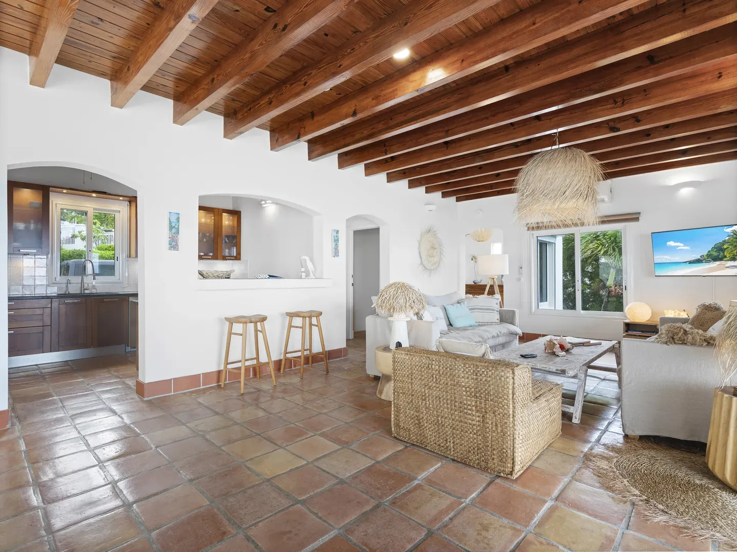 Open-concept living room and kitchen with terracotta tiles, exposed wood beams, wicker furniture, and outdoor greenery views
