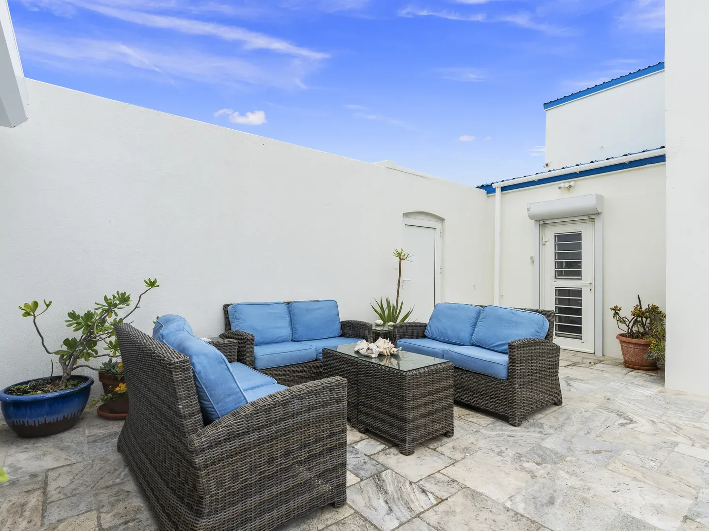 Villa outdoor terrace with wicker furniture, blue cushions, stone flooring, potted plants, and a clear blue sky