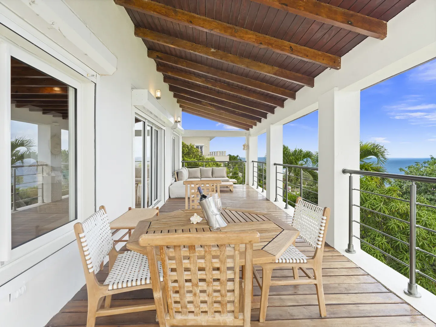 Covered wooden terrace with dining set, sofa, and ocean view through tropical greenery
