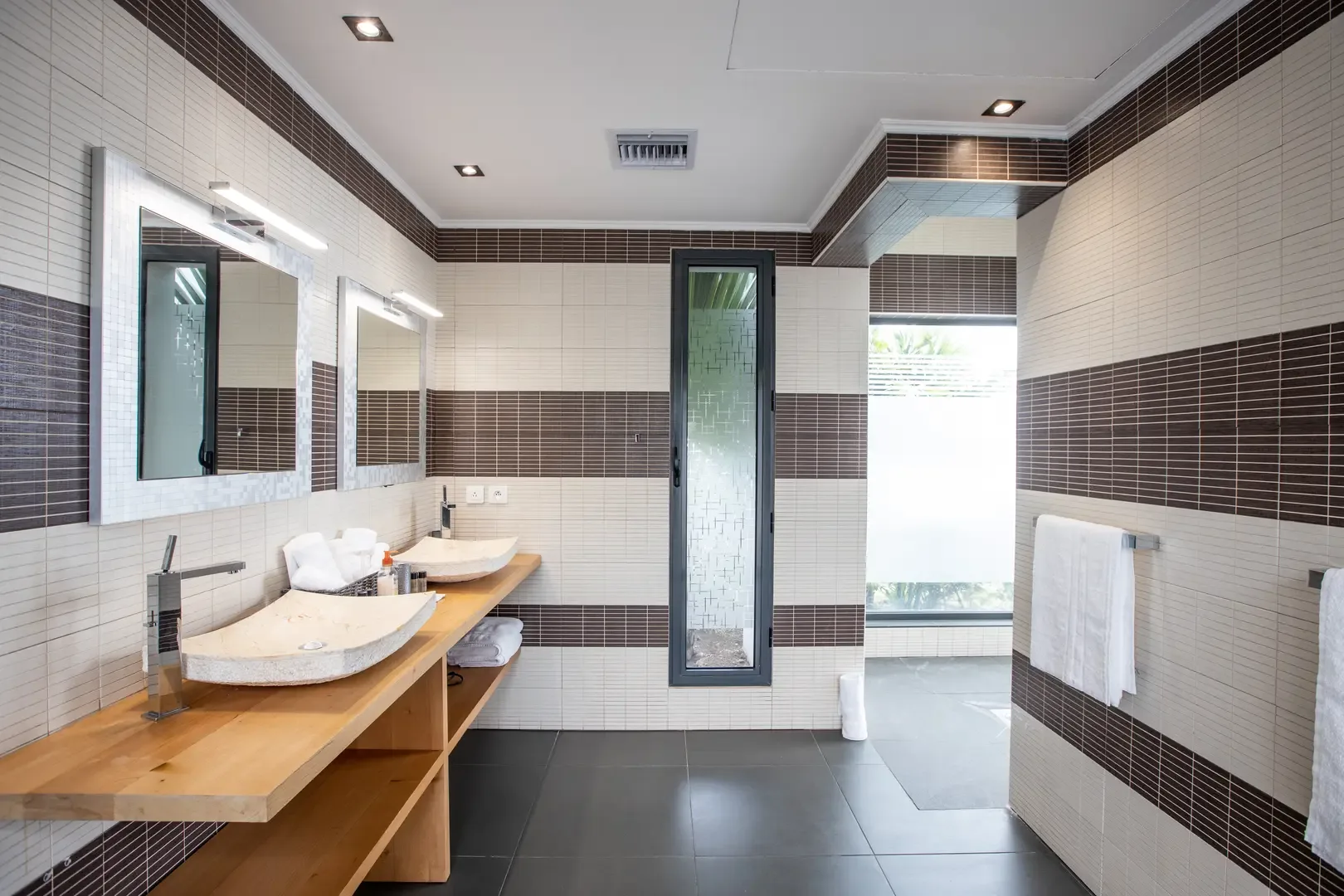 Modern luxury meets natural materials in this bathroom featuring double vessel sinks on a wooden floating vanity, integrated mirror lighting, and stylish brown and cream tiled walls.