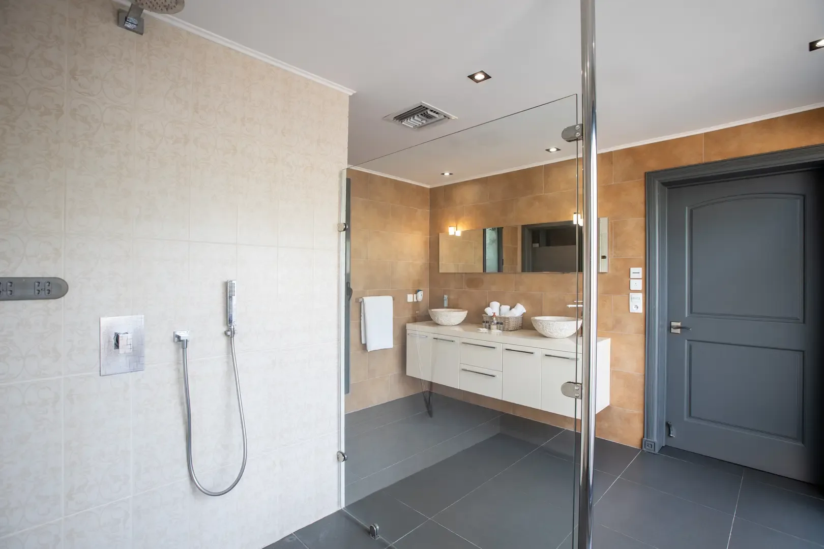 A glass-enclosed walk-in shower with rain showerhead stands beside the floating double vanity with vessel sinks, all set against dark gray tiled floors.