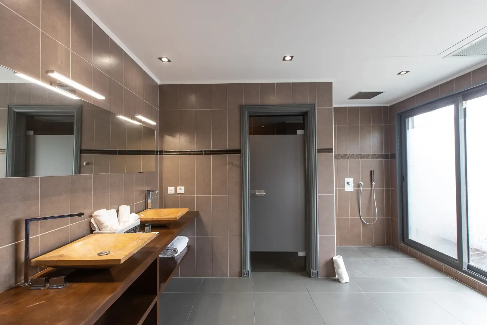 Natural light floods this modern bathroom through large windows, highlighting the dual vessel sinks, wooden vanity, and walk-in shower area.