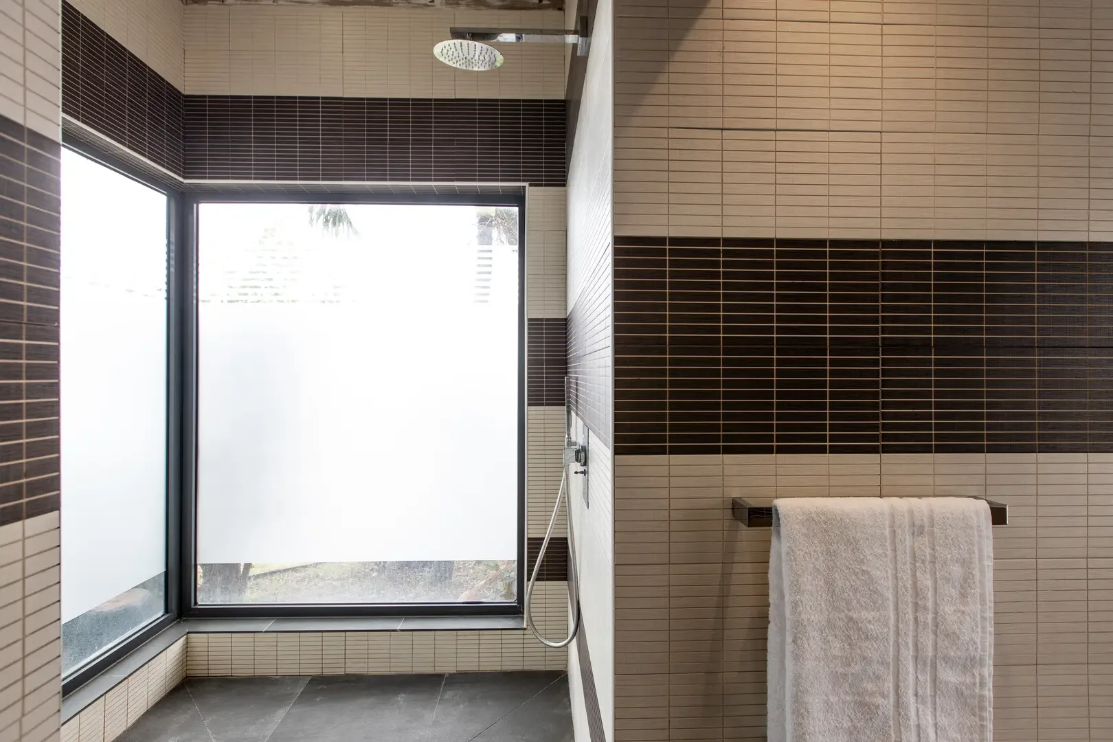 The modern shower area features a rainfall showerhead, frosted glass window for natural light, and dark slate flooring with beige and brown horizontal wall tiling.