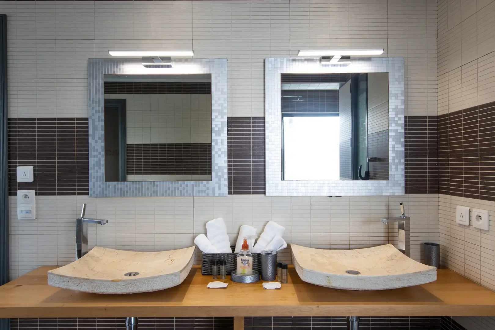 Double stone vessel sinks rest on a wooden vanity, while mosaic-framed mirrors with overhead lighting add elegance to the modern bathroom design.